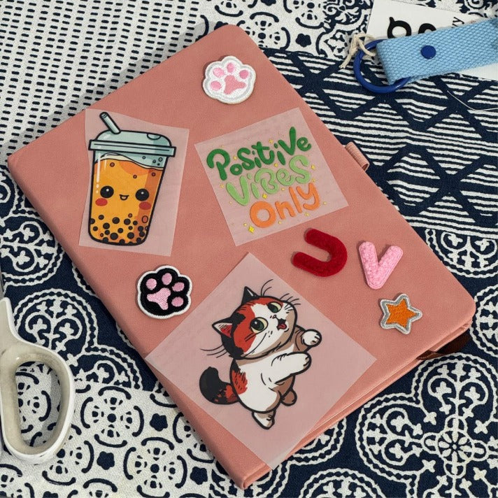 Close-up of a personalized pink A5 notebook decorated with custom stickers and patches including a boba tea, a cat, and "Positive Vibes Only" text.