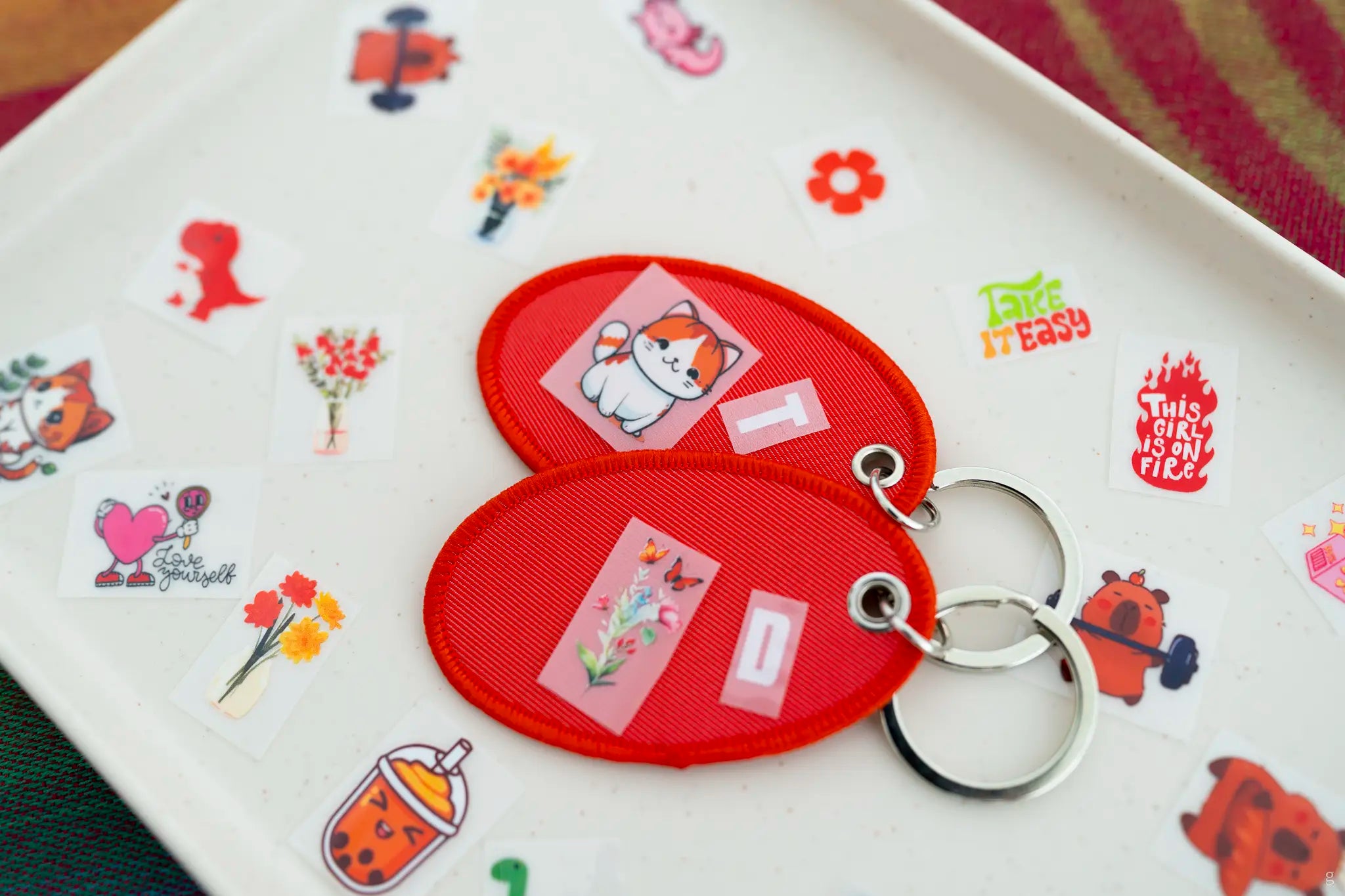 A pair of Red oval-shaped keychains with cartoon designs on a tray with printed patches from The Goodayy gifting studio located here in Singapore.