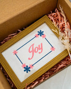 Medium wood frame featuring embroidered pink cursive text reading "Joy" inside a dotted border with blue and pink flowers, packaged in a box.