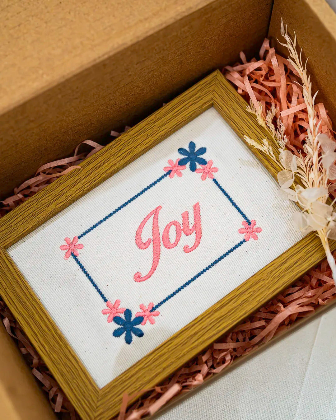 Medium wood frame featuring embroidered pink cursive text reading "Joy" inside a dotted border with blue and pink flowers, packaged in a box.