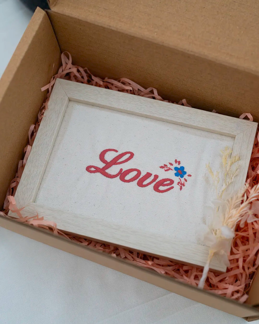 Embroidered white fabric frame with cursive pink text reading "Love" and small floral details, presented in a gift box with pink paper shred.