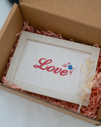 Embroidered white fabric frame with cursive pink text reading "Love" and small floral details, presented in a gift box with pink paper shred.