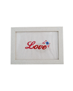 Light wood frame featuring red cursive embroidered text reading "Love" and a small floral detail on cream canvas.