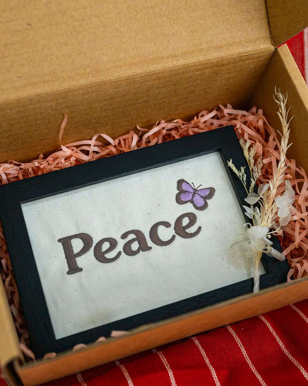 Black-framed embroidered artwork featuring the word "Peace" in dark thread and a purple butterfly on cream fabric, packaged in a gift box.