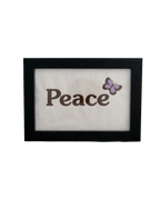 Black wood frame featuring the word "Peace" embroidered in dark thread with a purple butterfly on cream canvas.