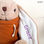 Close-up of custom embroidered name ‘Grace’ in purple on bunny ear of plush toy in orange hoodie