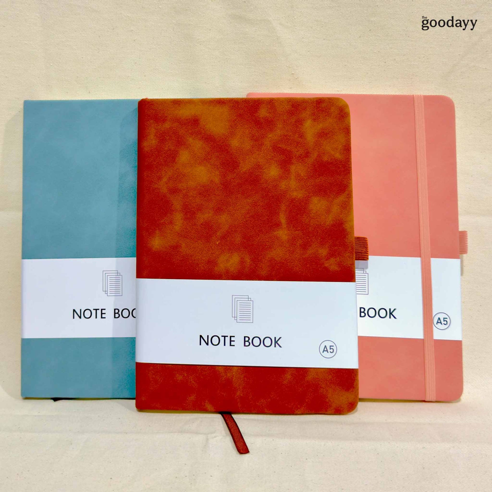 Three notebooks in blue, orange, and pink with 'the goodayy' branding on a beige background.
