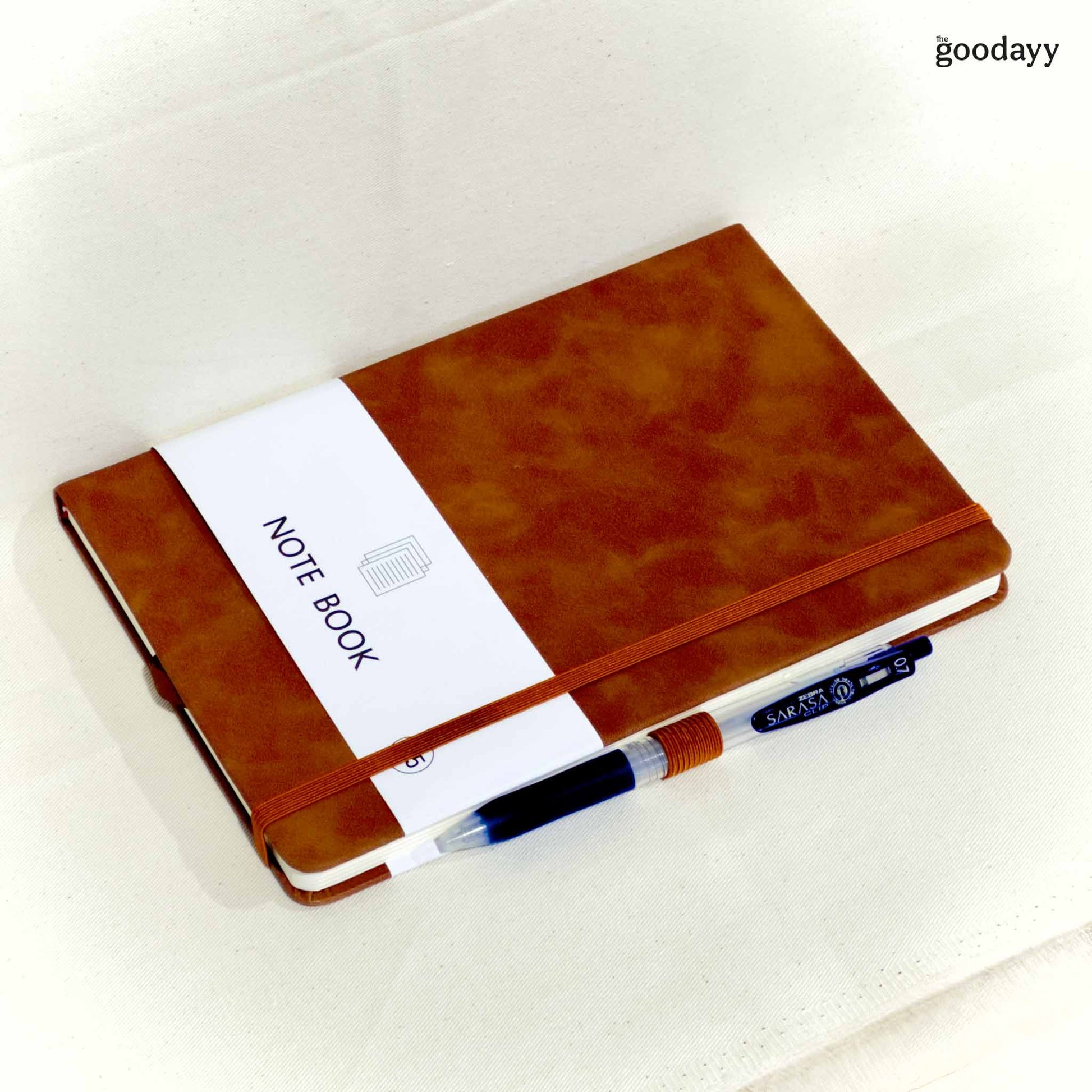 Brown notebook with a pen on a white background, branded 'the goodayy'.