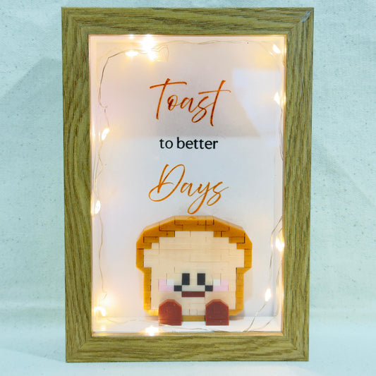 Decorative frame with toast-shaped lego and 'Toast to better days' text with fairy lights lighting up the frame on the inside.