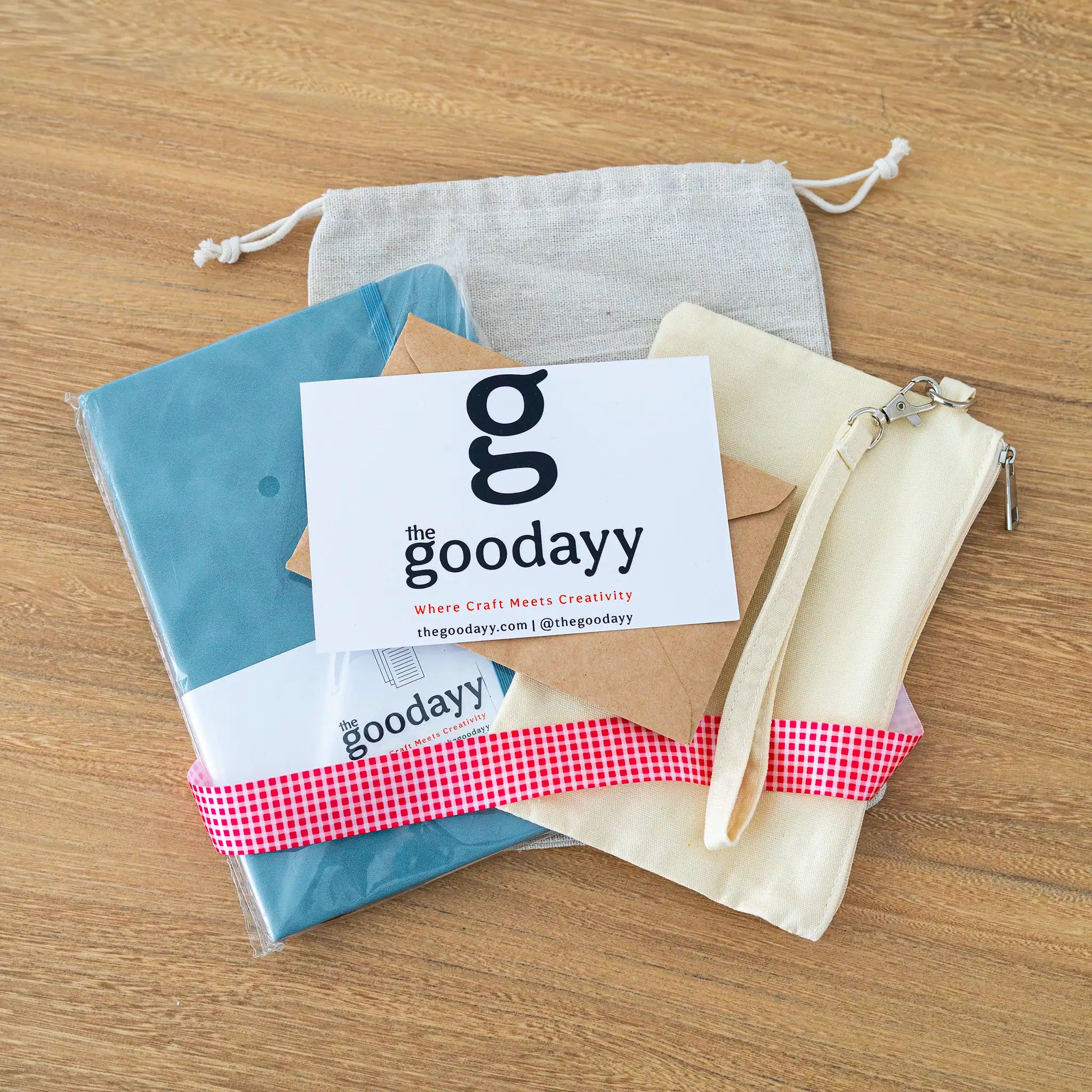 The Goodayy personalized gift bundle featuring a blue notebook, cream wristlet pouch, and canvas drawstring bag with a red gingham ribbon and greeting card.